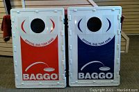 Two folded plastic bean toss boards side by side. One red with white print, one blue with white print. Each has a hole near top center for bean tossing.