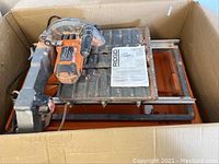 Ridgid wet tile saw in original box showing cutting table, motor housing, and instruction manual on top.