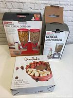 Photo of boxed Home Basics dual cereal dispenser, single cereal dispenser, and Choco Maker fondue melter tray on a white surface against a plain background.