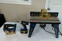 Image showing Ryobi cordless jig saw, cordless impact driver with battery, and router table grouped on a white surface.