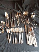 Photo showing assortment of silverplate flatware items including forks, knives, spoons, small pitcher, and candle holders laid on dark cloth.