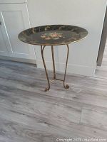 Full view of the ornate round black and gold metal side table showing three gold legs and floral design on top.