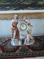 Full view of the porcelain clock with woman and two cherubs on a decorative base, showing scale and overall appearance.