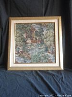Full view of tapestry art in gold wood frame and linen mat, showing detailed needlework depicting a riverside scene with people and birds.