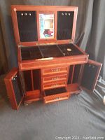 Full view of wooden jewelry chest opened showing top lid with mirror, black velvet panels with hooks, compartments, and side doors open with drawers below.