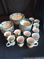 Overview of the dinnerware set arranged on black fabric, showing stacks of plates, bowls, and multiple mugs. Floral pattern visible with blue-green accents.