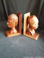 Pair of teak wood hand-carved bookends showing side profiles of human heads, placed on a black cloth background.
