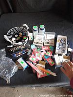 Wide view of all the assorted beauty items spread out on a dark surface including bowls, tweezers, eye shadows, manicure items, dental floss, and toe rings.