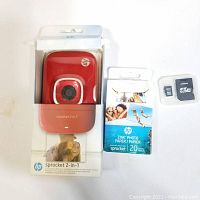 Device in original retail packaging with box, HP Sprocket 2-in-1 in red, 20 sheets of HP ZINK photo paper for printing small photos, and SanDisk 8GB microSD card.