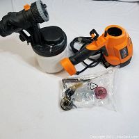 Electric spray gun with orange handle and black nozzle alongside white paint container; accessories bag visible.