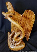 Front and side views of the hand carved wooden eagle sculpture holding a snake. Shows the detailed carving of feathers, the snake wrapping around the talons and being held in the beak, and the natural wood finish. The eagle has a broken tip on the beak.