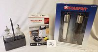 Photo showing boxed KitchenAid 8-piece food prep set, boxed Starfrit salt and pepper grinders, and glass oil and vinegar bottles in metal stand.
