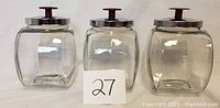 Front view of three clear glass canisters with silver lids and maroon knobs, placed side by side with a lot number card in front.