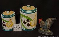 Photo showing two hand-painted ceramic canisters with olive and garlic designs alongside painted metal rooster doorstop, on black background.