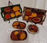 Five hand painted platters with colorful fruit designs arranged on floor, two larger rectangular with handles and three smaller oval/rectangular platters.