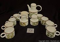 Photo showing entire twelve piece tea set arranged with eight mugs, two creamers, one sugar bowl, and one teapot. Pieces have white base with green and yellow floral pattern and brown rim.