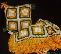 Handmade crochet afghan blanket in white, yellow, and green yarn with yellow fringe, paired with matching square pillow with tassels, shown on black background.