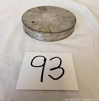 Closed round antique tin photographed from above on white background with lot number 93 card next to it