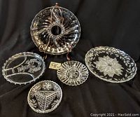 Five glass platters of varying sizes arranged on a black fabric background. Visible intricate floral and geometric etched designs on clear glass surfaces.