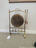 Front view of hammered brass gong hanging from arched brass frame with mallet at right.
