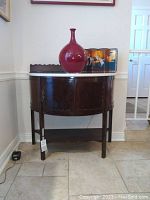 Front view of antique wooden semi-circular entryway cabinet with white marble top, decorative carved wooden body, and metal handles.