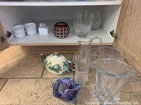 Full view of lower cabinet shelf and floor showing ceramic teapot, clear glass carafe, clear crystal and fluted glass vases, hand-blown glass bowl in front.
