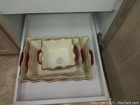Set of 3 ceramic baking dishes nested inside each other, cream-colored with red handles and scalloped edges, showing Sur La Table branding on the bottom.