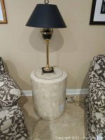 The travertine pedestal base with cylindrical shape, natural stone finish, positioned in between patterned fabric chairs with lamp placed on top.