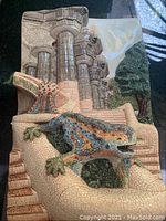 Front and side angles showing the colorful ceramic lizard sculpture with mosaic style, stone column backdrop, and green tree.