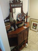 Wooden dresser with decorative items including tall floral vase, brass container, decorative plates, and other ceramic and wooden items.