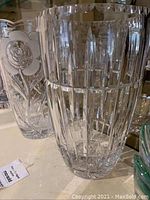 Photo showing a tall crystal vase with vertical panel cuts and another crystal vase with a floral relief pattern behind it.