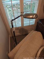 Photo showing floor lamp placed behind beige armchair near window with blinds, capturing adjustable stainless steel shade and wood arm components.