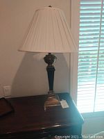 Black table lamp on a dark wood table near window blinds, showing the entire lamp and pleated white shade