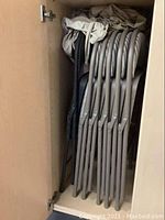 Five grey folding chairs stacked and stored inside a cabinet, showing their metal frames and fabric backs and seats, with some wear visible.