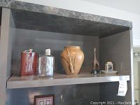Photo showing two hip flasks, beige ceramic vase, small wooden giraffe figurine on a metal shelf