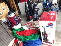 Snowman figurine with dark blue and red winter clothing, deflated blow-up decorations, box of candy cane ornaments, Hoover vacuum box, vacuum cleaners in background.