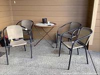 Photo of four black metal framed outdoor chairs with beige canvas seats and backs around a small round folding table with glass top and plastic wicker edge, placed on a patio with brown siding.