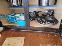 Cabinet shelf with turquoise nonstick saucepans and sauté pans, several stainless steel pots, and nonstick frying pans.