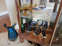 View showing art glass vases, sculptures on bottom shelf, amber bowls on glass shelf, and small blue perfume bottle.