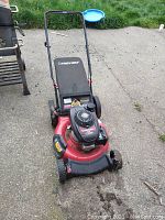 Front-left angle view of the red Troy-Bilt lawn mower on concrete with wheels and grass catcher bag visible, showing some dirt buildup.