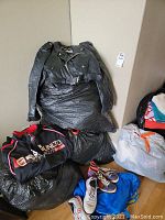 Photo showing a black leather biker jacket on top of several black plastic bags with other clothing items, and two pairs of shoes on the floor.