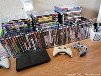 Shows a black PlayStation 2 console, three controllers (two white, one translucent blue and grey), and a large collection of PlayStation 2 game discs in cases arranged on a wooden surface.