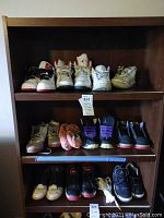 Photo showing multiple pairs of men's shoes arranged on three shelves of a wooden shelving unit. Includes white, black, grey, and multicolor basketball and casual shoes. Condition used, some dirty and worn.