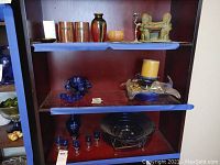 Wooden shelf with various decorative pieces including tall wooden glasses, various vases, a sculpted figure on top shelf; blue art glass candle holder with candle on middle shelf; glass bowl on metal stand and small blue glasses on bottom shelf.