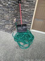 Green garden hose coiled on ground with black plastic shovel with red handle resting on top, near stone wall and outdoor faucet
