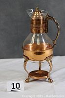 Front view of vintage glass coffee carafe with copper collar, ornate handle, and copper warming stand beneath.