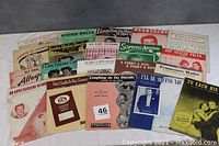 Lot of 20 vintage sheet music pieces laid out overlapping, showing variety of colorful and graphic covers with some visible titles and images.