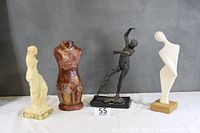 Group photo of four statues on white surface against gray background: cream Venus De Milo on marble, reddish-brown dress form, black dancing figure, modern white abstract figure on wood base