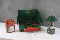 Photo showing green Coleman two-burner camping stove open with wind guards, red rusty Coleman fuel can, and Coleman green lantern with wire handle on a grey background.