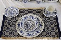 Dinnerware plates, bowls, cup, and saucers arranged on a woven blue and white placemat.